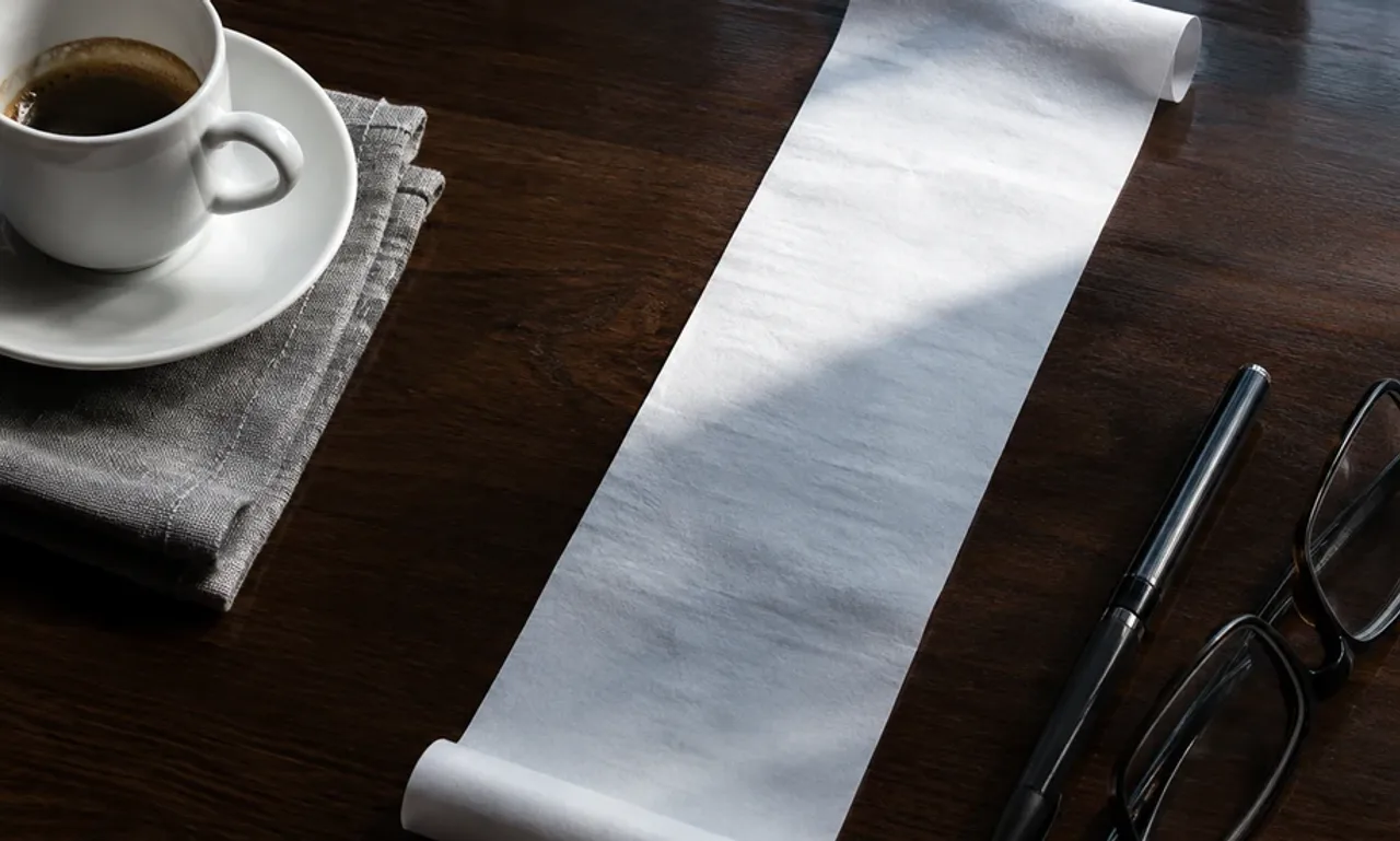 A long blank paper receipt on a dark walnut desk, half lit by a shaft of cool daylight and half in shadow, with an espresso cup, a pen and folded reading glasses beside it.